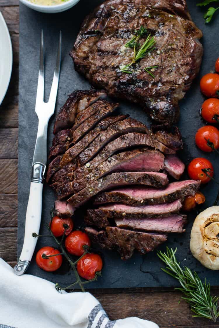 Sliced grilled dry aged rib eye steaks resting on a slate platter with grilled tomatoes, garlic.