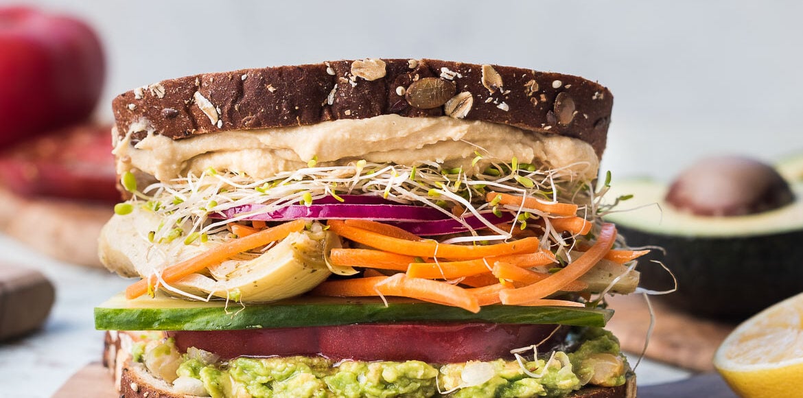 Avocado Veggie Sandwich with hummus, alfalfa sprouts, pickled onions, carrots, artichoke, cucumber, and tomato on a wooden board.
