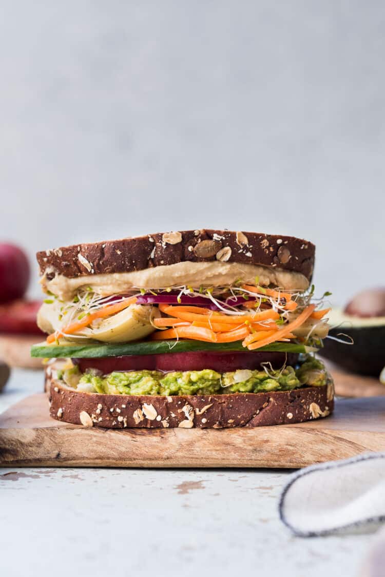 Avocado Veggie Sandwich with hummus, alfalfa sprouts, pickled onions, carrots, artichoke, cucumber, and tomato on a wooden board.