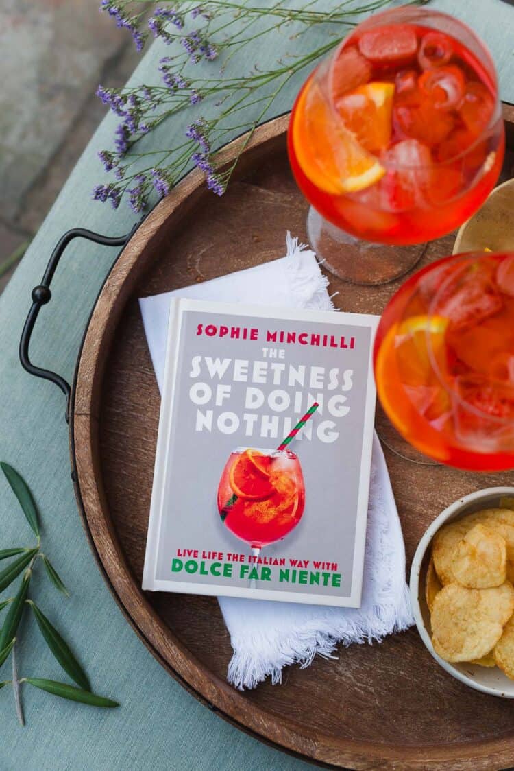The Sweetness of Doing Nothing, by Sophie Minchilli, on a tray, with glasses of Aperol Spritz.