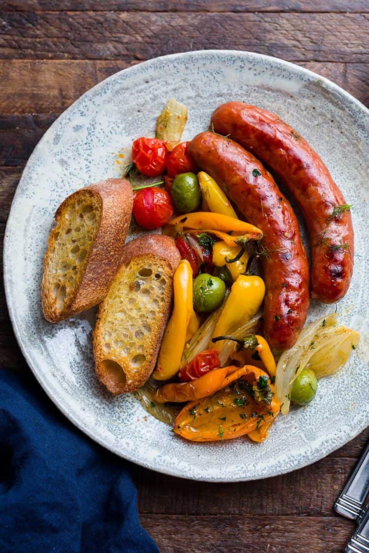 Plate with sheet pan-roasted sausages, peppers, onion, fennel, tomato, and olives, with toasted baguette.