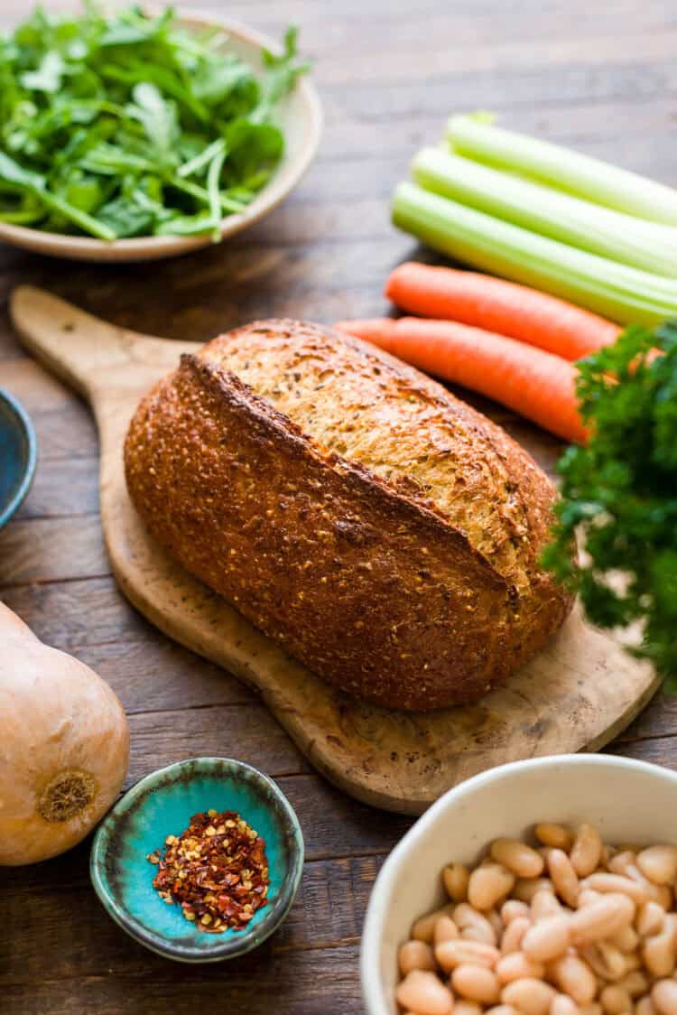 Whole grain bread on a cutting board with ingredients for a Ribollita (Tuscan-style Bread Soup) recipe.