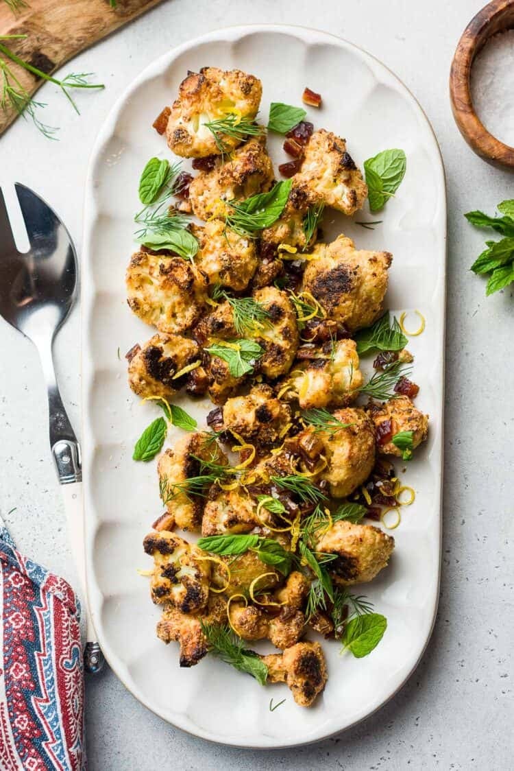 Tahini-Charred Cauliflower garnished with dates, mint, dill, and lemon zest, on a serving platter.