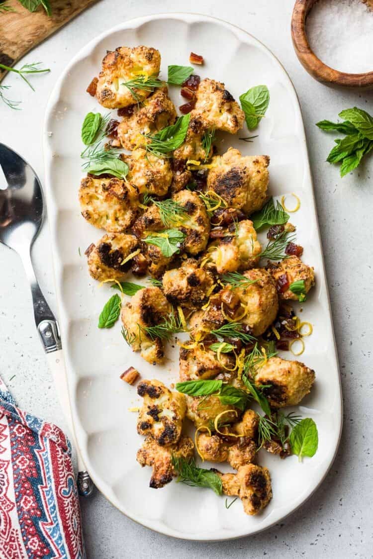 Tahini-Charred Cauliflower garnished with dates, mint, dill, and lemon zest, on a serving platter.