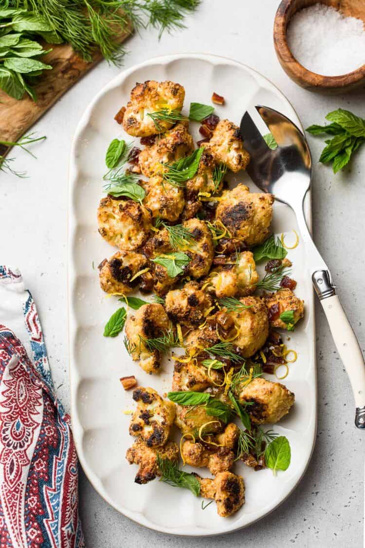 Tahini-Charred Cauliflower garnished with dates, mint, dill, and lemon zest, on a serving platter.