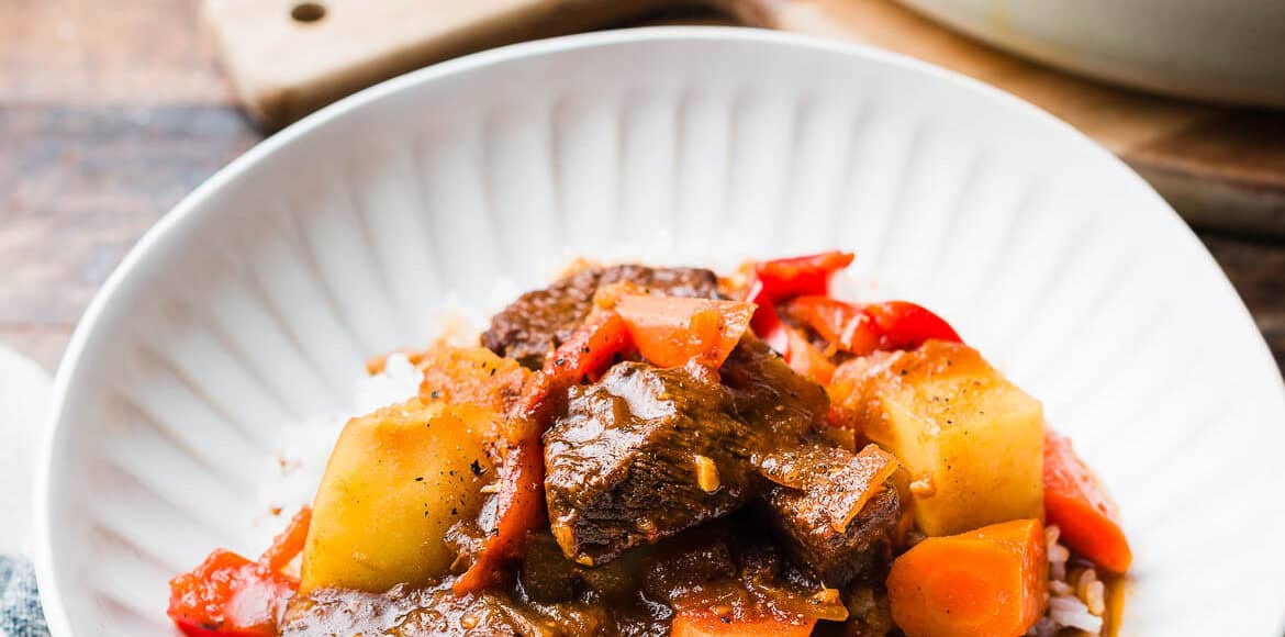 Mechado (Filipino Beef Stew) served over rice in a white bowl, with a large pot of mechado in the background.