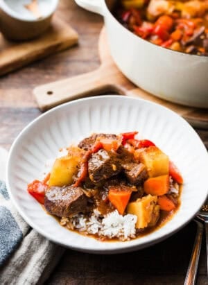 Mechado (Filipino Beef Stew) served over rice in a white bowl, with a large pot of mechado in the background.