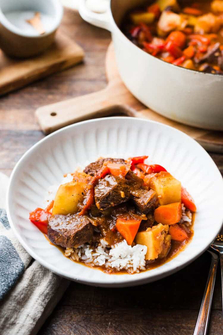 Mechado (Filipino Beef Stew) served over rice in a white bowl, with a large pot of mechado in the background.